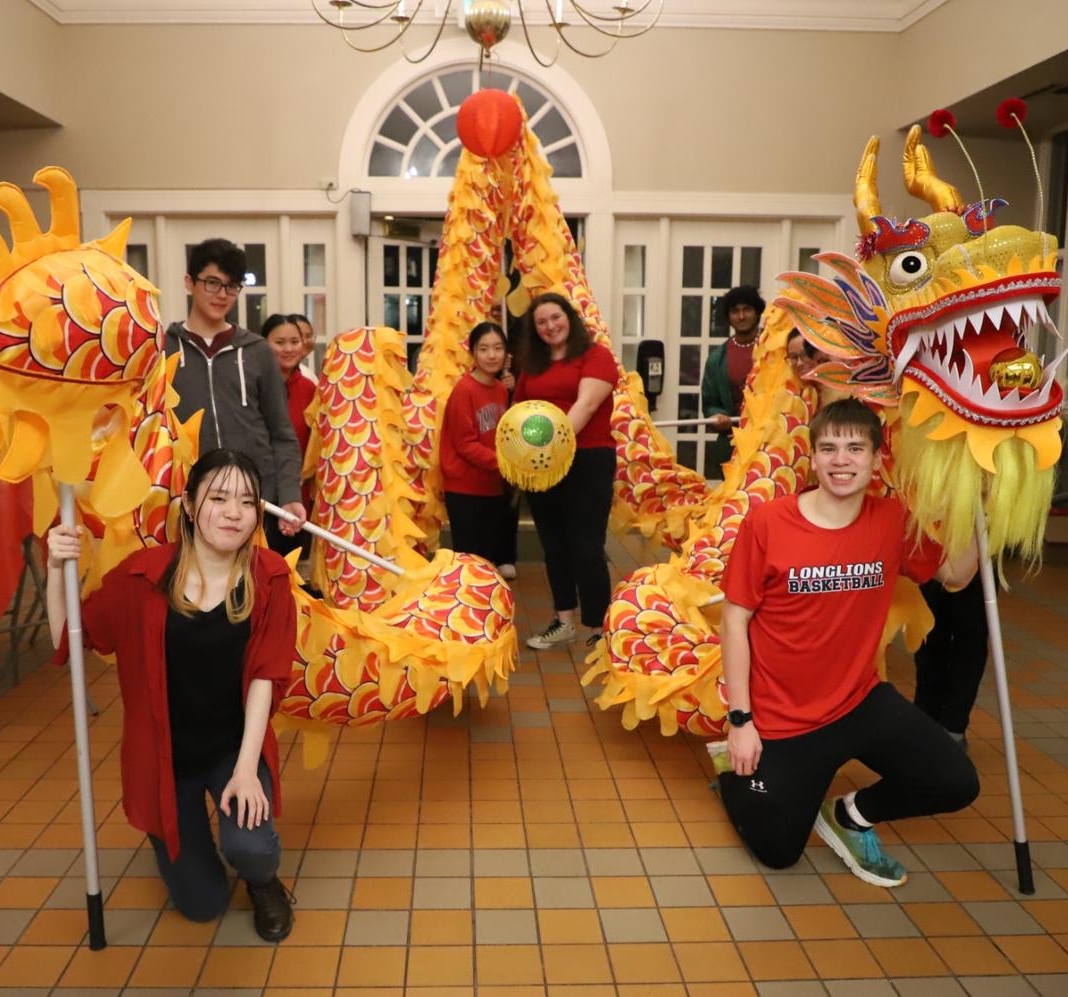 A group of people pose indoors holding a yellow and orange dragon costume, with a tiled floor and arched windows in the background.
