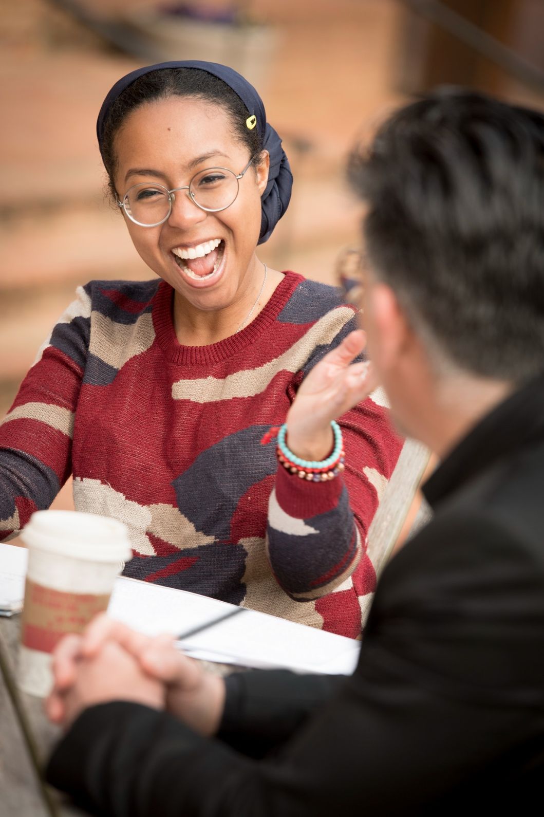 A woman wearing glasses and a patterned sweater is smiling and gesturing while talking to a man across a table with coffee cups and papers.