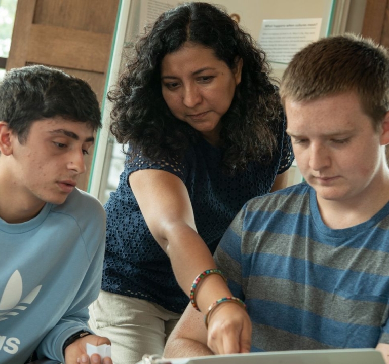 A woman points at a laptop screen while two young men look on, appearing focused and engaged in a discussion or lesson.
