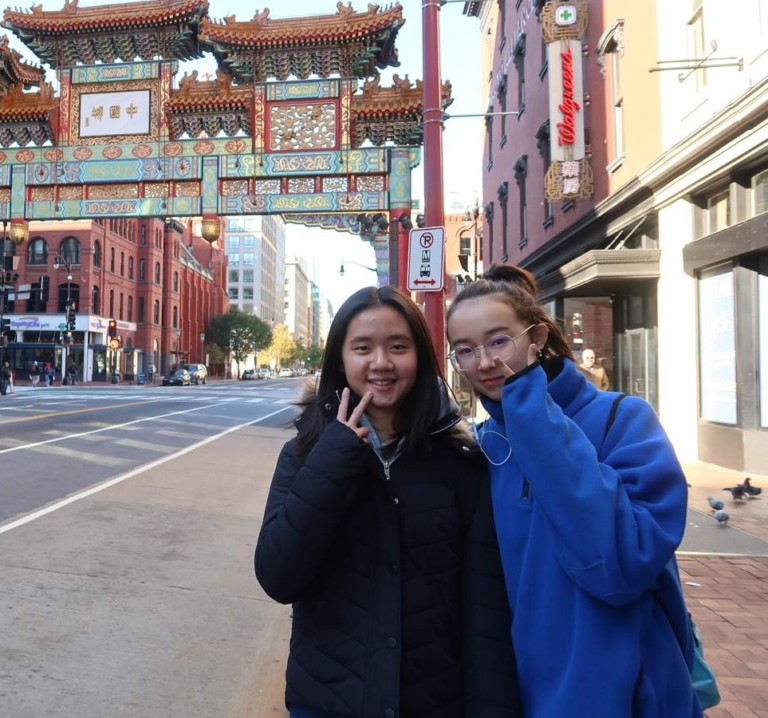 Two young women stand on a city street in front of a traditional 中国 archway, with buildings, a bus, and Walgreens visible in the background.