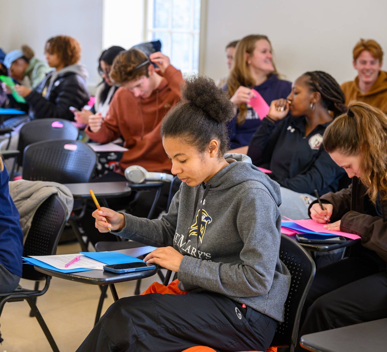 Students sit at desks in a classroom, writing on colorful paper and engaging in an activity, with some looking focused and others interacting.