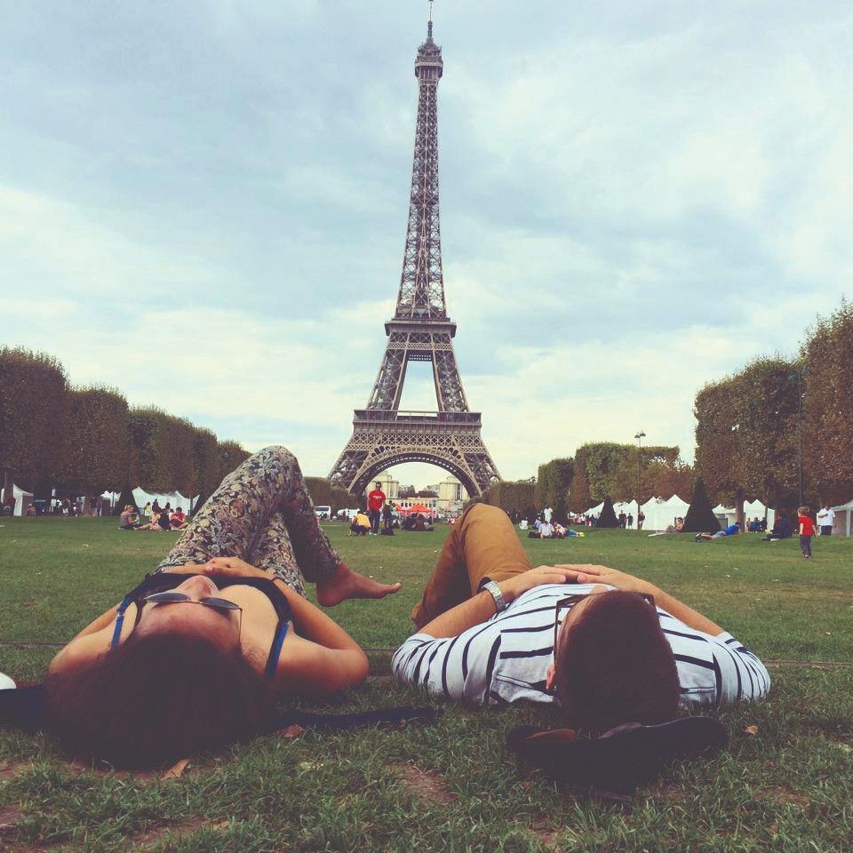 Two people lie on the grass facing the Eiffel Tower in Paris, with trees and other visitors in the background under a cloudy sky.