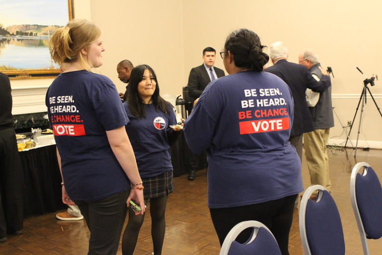 Three women in matching “Be Seen. Be Heard. Be Change. Vote” shirts talk in a conference room while others mingle in the background.