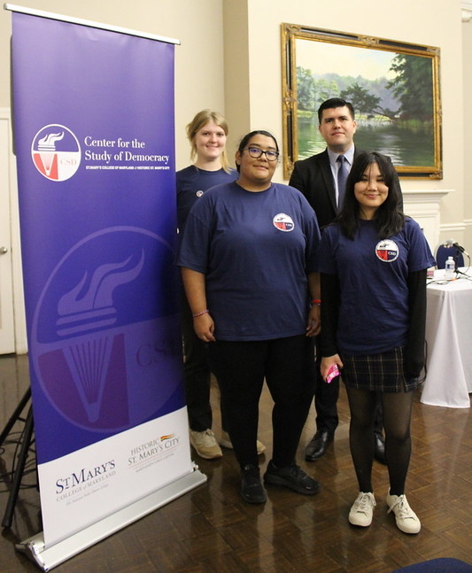 Four people stand indoors next to a 民主研究中心 sign; three wear matching blue shirts, and one is in a suit.