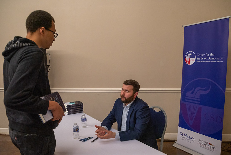 A man seated at a table speaks with a standing attendee at a book signing event hosted by the Center for the Study of Democracy.