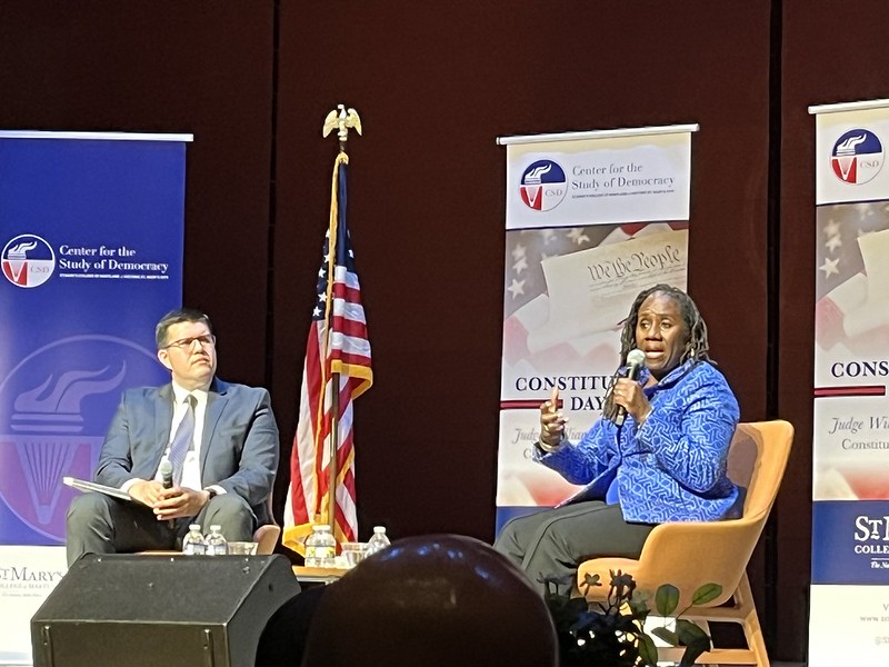 Two people are seated on stage having a discussion, with an American flag and banners for the 民主研究中心 in the background.