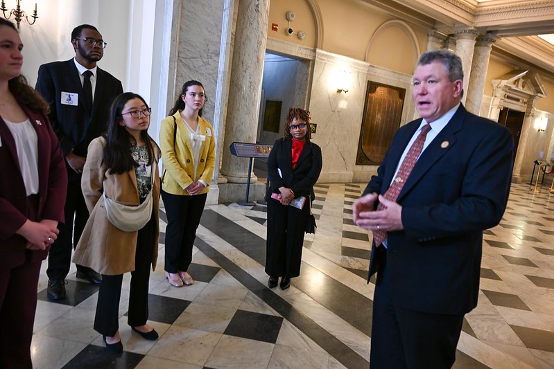 A man in a suit speaks to a group of people standing in a marble-floored hallway with columns and ornate lighting.
