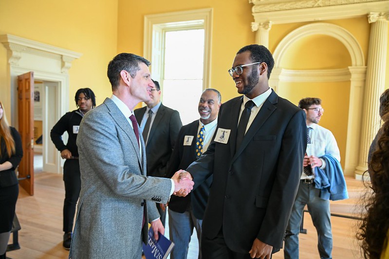 Two men in suits shake hands and smile in a formal room, while others stand and observe in the background.