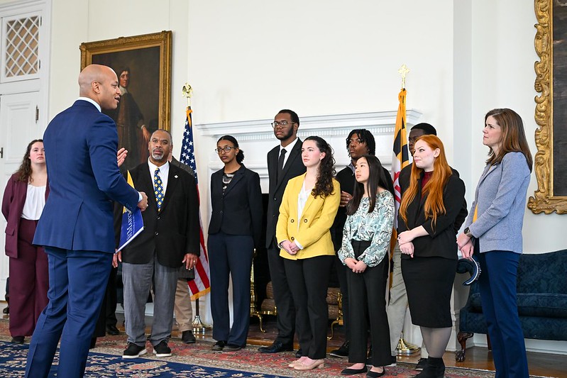 A man in a suit speaks to a group of people standing in a formal room with flags, a fireplace, and a large portrait in the background.