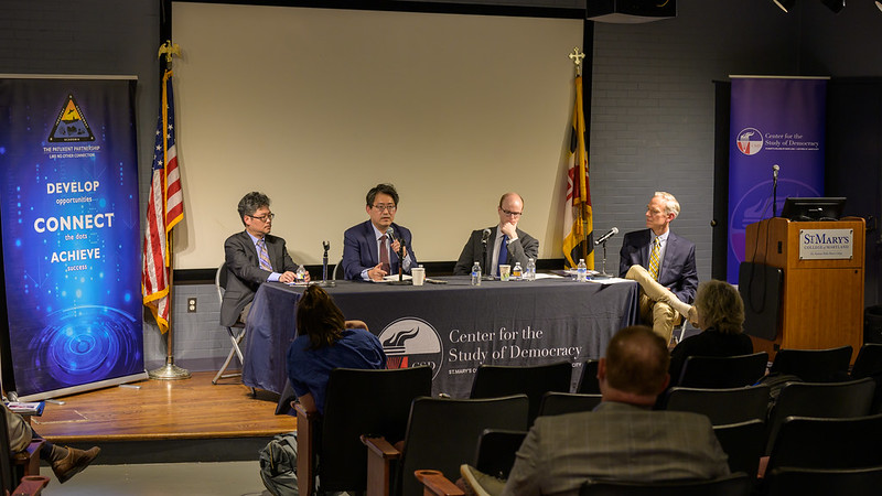 Four men sit at a panel discussion table in front of an audience, with banners, a projector screen, and flags behind them at the 民主研究中心 event.