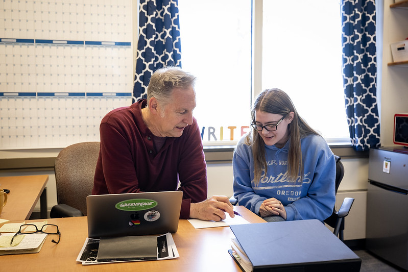 A man and a young woman sit at a desk, reviewing papers together in an office with a laptop, calendar, and window in the background.