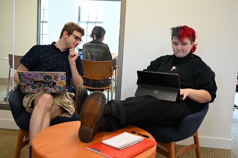 Two people sit in chairs with laptops; one looks at the other while they work. A notebook and pen are on a small table in front of them.