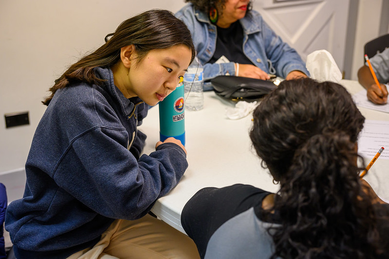 Two people sit at a table having a conversation while others are also seated nearby, with papers and a water bottle visible on the table.