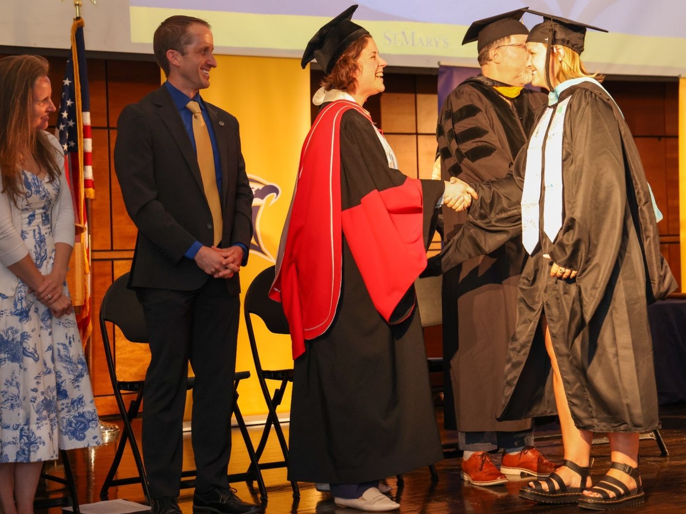 A graduate in academic regalia shakes hands with a faculty member on stage during a graduation ceremony, with others standing and watching.