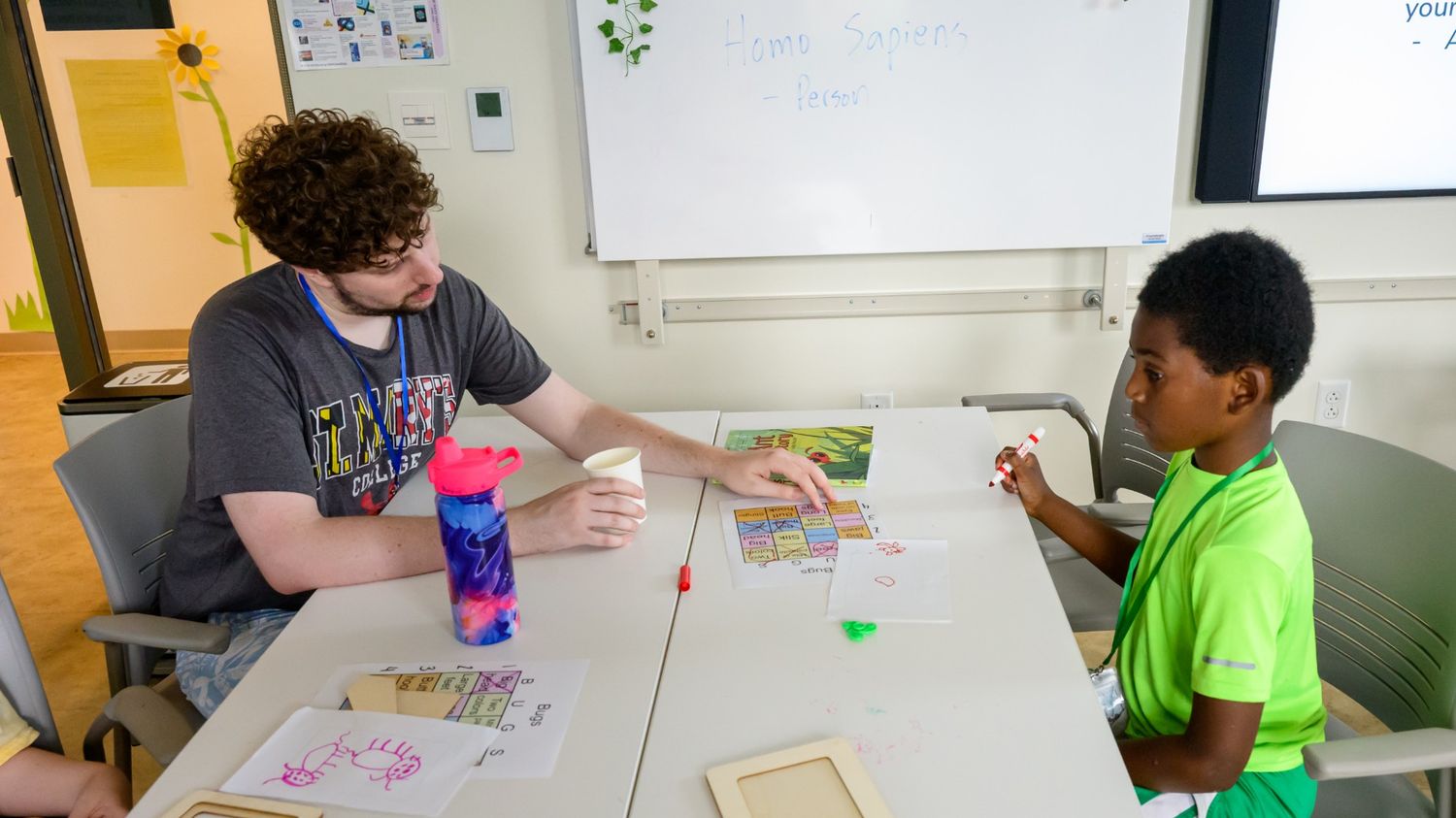 An adult and a child sit at a table doing arts and crafts activities with papers, markers, and colored objects in a classroom setting.