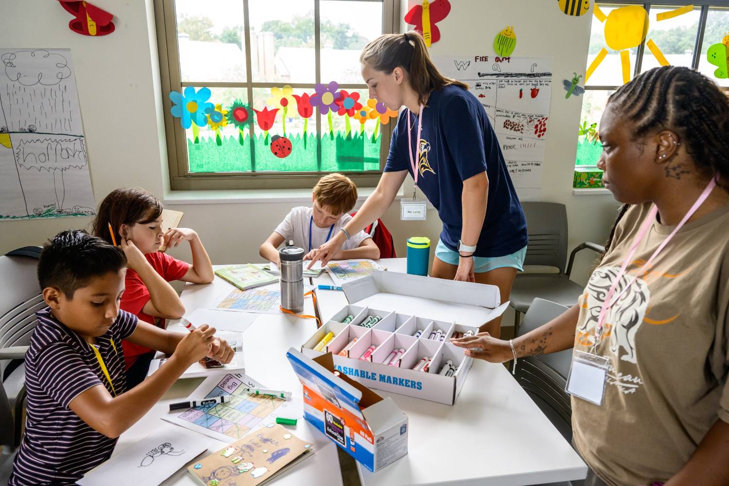 Four children sit at a table working on crafts with markers while two adults supervise in a brightly decorated classroom.