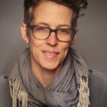 Person with short, styled hair and glasses, wearing a gray scarf and dark shirt, smiles against a neutral background.