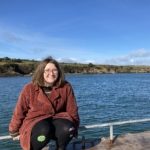 A person with glasses and medium-length hair sits on a rail by a waterfront. They are wearing a red coat and black pants, smiling with a scenic backdrop of blue water and a hilly landscape under a clear blue sky.