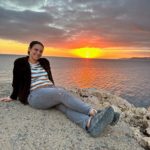 A person sitting on a rocky surface by the water, smiling at the camera. They are wearing a striped shirt, dark jacket, and jeans. In the background, there's a vivid sunset with orange and pink hues over the ocean horizon and a partially cloudy sky.