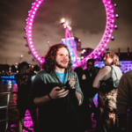 A person with curly hair holds a smartphone and smiles outdoors at night. A large, illuminated Ferris wheel, lit in pink, is visible in the background. Several other people are also visible in the scene.