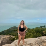 A woman in a black tank top and brown shorts stands on a rocky surface with her hand on her hip. She smiles at the camera with a backdrop of lush greenery and a vast ocean under a cloudy sky.
