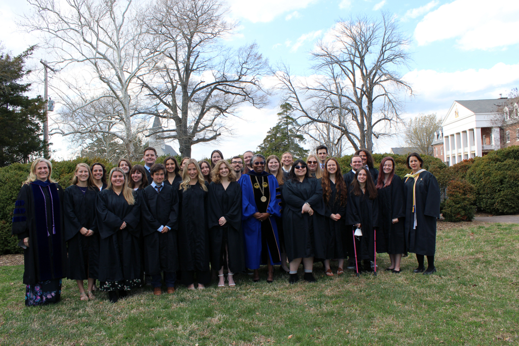 A group of people in academic robes stand together on a grassy lawn, posing for a group photo. Trees and a building are visible in the background. The sky is partly cloudy.