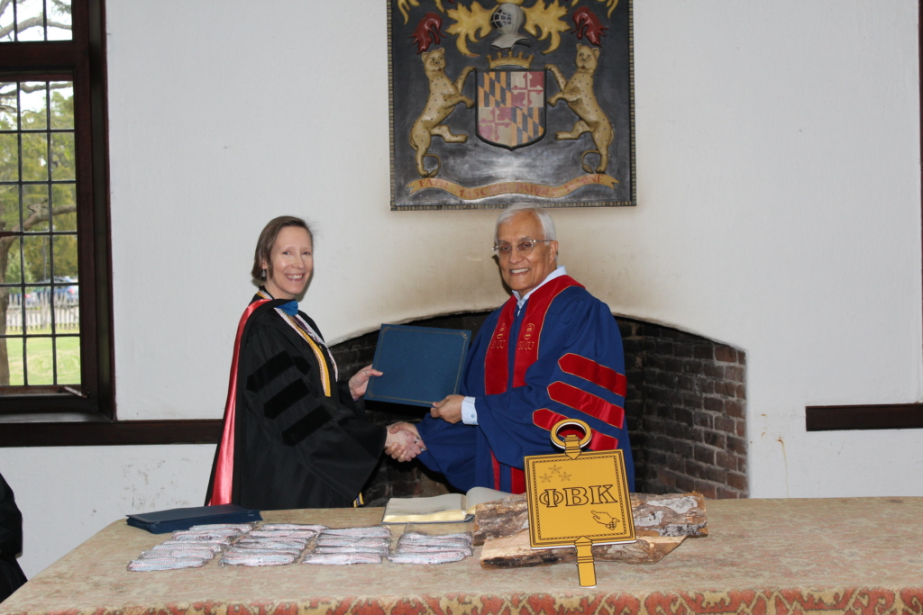 A woman and a man in academic robes are smiling and shaking hands during a formal ceremony. There's a large key with Greek letters on a table in front of them and a coat of arms on the wall behind them.