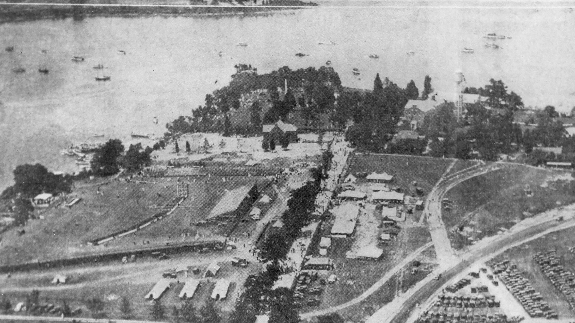 Aerial view of a coastal area with a small peninsula. There are numerous tents and structures on open grassy fields, surrounded by trees. Boats are anchored in the water nearby. A road and several parked cars are visible on the right.