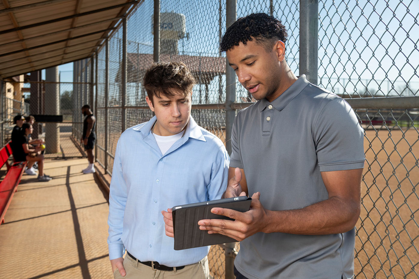 A student in the sport and recreation program speaks with another in a baseball dugout while holding up an iPad with scheduling information on it. 