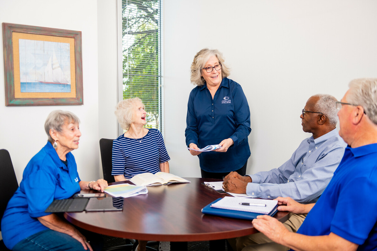 Five older adults sit around a small round table in a bright office, engaged in discussion. One woman stands, handing out papers, while others listen and smile with notebooks and tablets open.